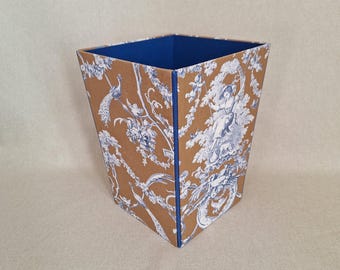 May include: A square wastebasket with a brown and white toile pattern featuring figures, trees, and peacocks. The interior is blue, and the edges are trimmed with blue. A decorative and functional item.