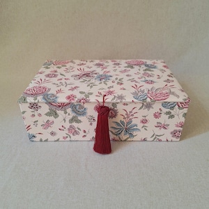 Pode incluir: Uma caixa de armazenamento retangular com uma base de cor creme apresentando um padrão floral repetitivo em tons de rosa, azul e verde. A tampa é fixada com uma borla decorativa vermelho-escura pendurada no centro da parte frontal.