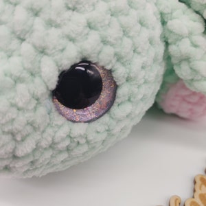 May include: Close-up of a green crocheted stuffed animal with a sparkly purple and black eye.