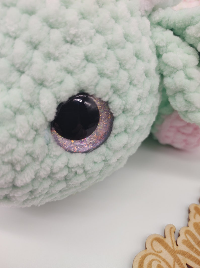 May include: Close-up of a green crocheted stuffed animal with a sparkly purple and black eye.