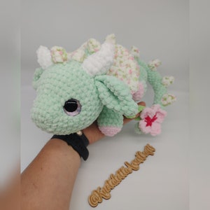 May include: A green crocheted dragon with a white horn and pink accents. The dragon has a floral pattern on its back and a pink flower on its tail. The dragon is sitting on a hand.