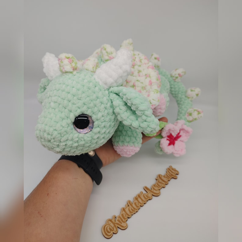 May include: A green crocheted dragon with a white horn and pink accents. The dragon has a floral pattern on its back and a pink flower on its tail. The dragon is sitting on a hand.