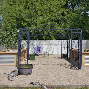 May include: A black metal garden arbor with a white picket fence in the background. Two raised garden beds are on either side of the arbor, each with a wooden frame and metal siding. A purple folding chair and a small table are inside the arbor.