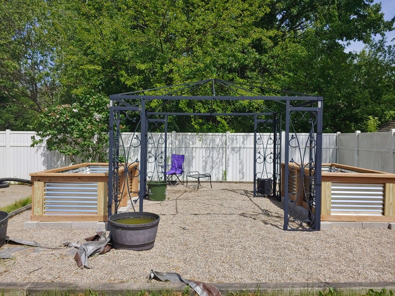 May include: A black metal garden arbor with a white picket fence in the background. Two raised garden beds are on either side of the arbor, each with a wooden frame and metal siding. A purple folding chair and a small table are inside the arbor.