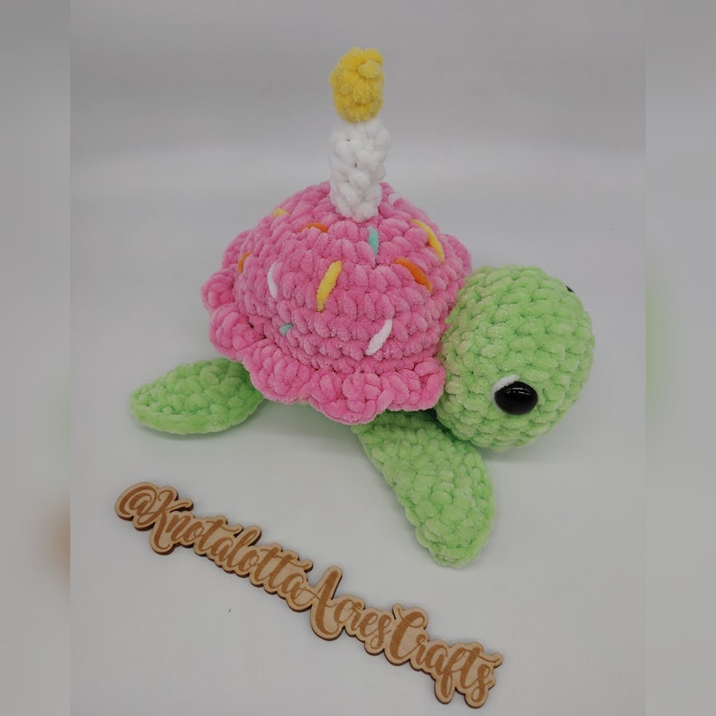 K&ouml;nnte beinhalten: Eine handgefertigte H&auml;kelschildkr&ouml;te mit einem rosa Cupcake-Panzer, verziert mit bunten Streuseln und einer wei&szlig;en Kerze mit gelber Flamme. Die Schildkr&ouml;te hat gr&uuml;ne Flossen und einen gr&uuml;nen Kopf sowie schwarze Augen. Das Holzschild sagt "@Knotalotta Acres Crafts".