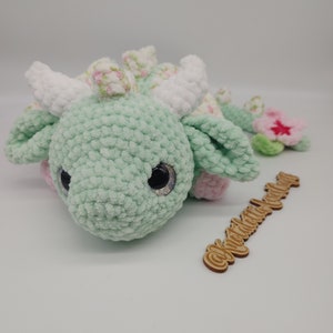 May include: A green crocheted dragon plush toy with white horns and pink accents. The dragon has large black eyes with glitter accents and a pink flower on its tail. The toy is lying on a white surface with a wooden sign that says "@knitcrochet_world".