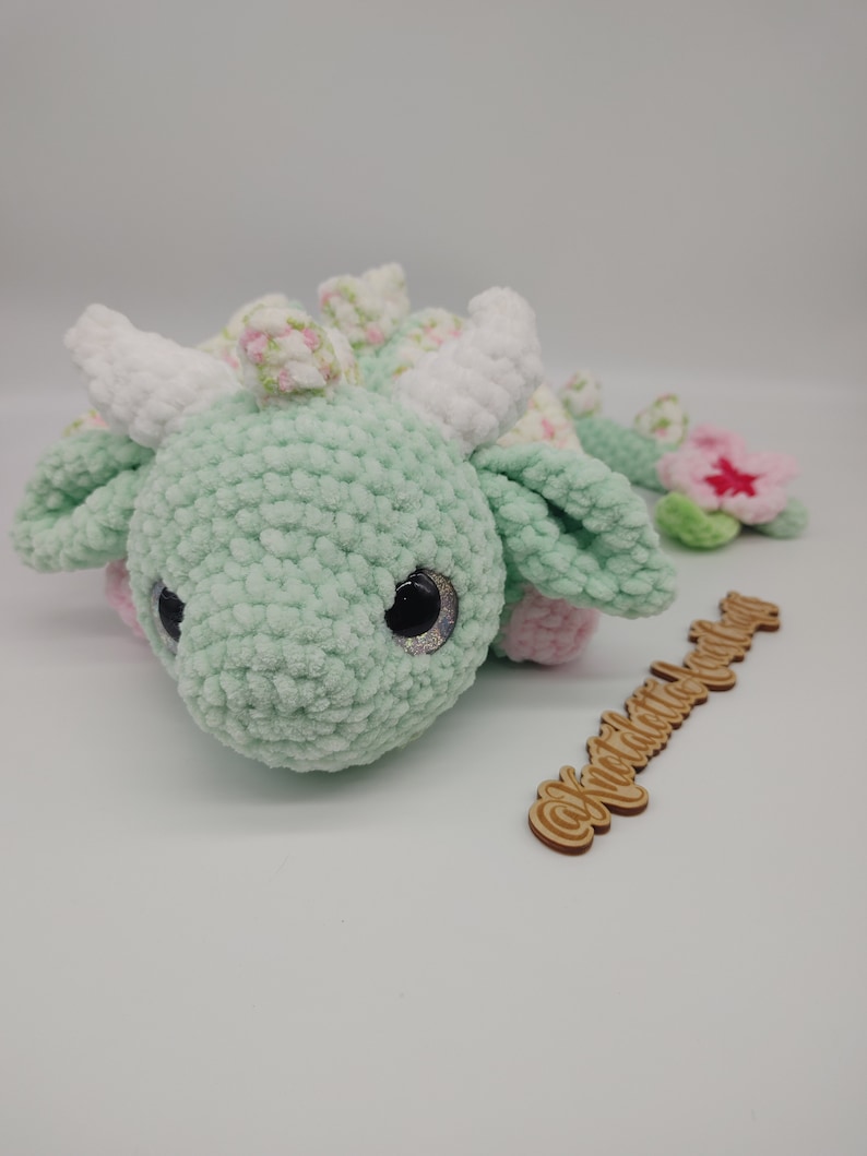 May include: A green crocheted dragon plush toy with white horns and pink accents. The dragon has large black eyes with glitter accents and a pink flower on its tail. The toy is lying on a white surface with a wooden sign that says "@knitcrochet_world".