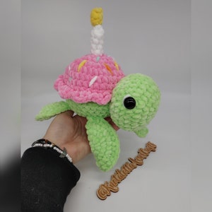 K&ouml;nnte beinhalten: Eine gr&uuml;ne Pl&uuml;schschildkr&ouml;te mit einer rosa Cupcake-Schale und einer wei&szlig;en und gelben Kerze. Die Schildkr&ouml;te hat schwarze Augen und ist aus weichem, strukturiertem Garn gefertigt. Die Cupcake-Schale ist mit bunten Streuseln verziert.