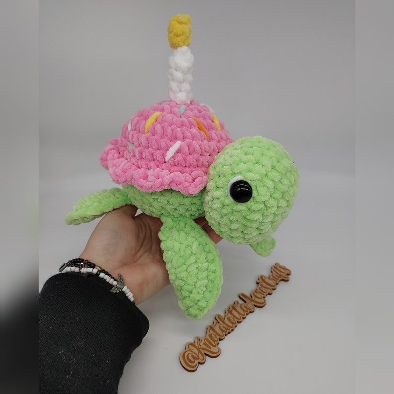 K&ouml;nnte beinhalten: Eine gr&uuml;ne Pl&uuml;schschildkr&ouml;te mit einer rosa Cupcake-Schale und einer wei&szlig;en und gelben Kerze. Die Schildkr&ouml;te hat schwarze Augen und ist aus weichem, strukturiertem Garn gefertigt. Die Cupcake-Schale ist mit bunten Streuseln verziert.