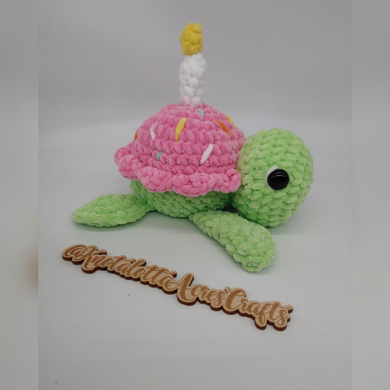 K&ouml;nnte beinhalten: Eine handgefertigte H&auml;kelschildkr&ouml;te mit einem rosa Cupcake-Panzer, verziert mit Streuseln und einer wei&szlig;en Kerze mit gelber Flamme. Die Schildkr&ouml;te ist hellgr&uuml;n mit schwarzen Augen. Das Holzschild sagt "Kristalotta Acres Crafts".