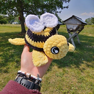 May include: A crocheted bee plush toy with yellow and black stripes, white wings, and black eyes. The bee is being held in a hand with bracelets.
