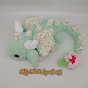 May include: A crocheted green dragon with a floral pattern on its wings and a pink flower on its tail. The dragon has white horns and a white belly. The dragon is lying on its side with its head turned to the left. The dragon is made of yarn and has a soft, cuddly texture.