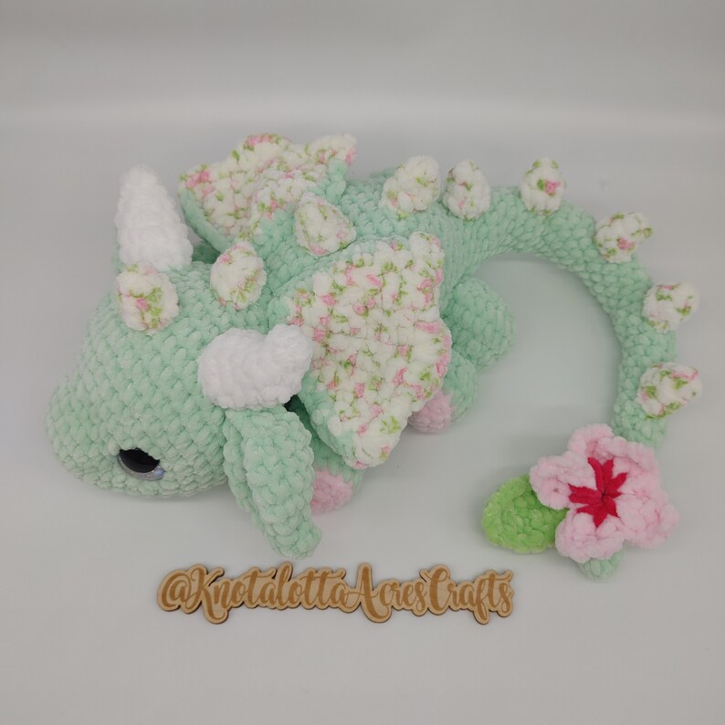 May include: A crocheted green dragon with a floral pattern on its wings and a pink flower on its tail. The dragon has white horns and a white belly. The dragon is lying on its side with its head turned to the left. The dragon is made of yarn and has a soft, cuddly texture.