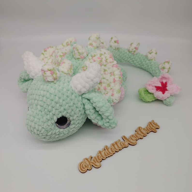 May include: A green crocheted dragon with a white crocheted horn and pink floral accents. The dragon has a long tail with a pink flower at the end. The dragon is lying on its side with its head resting on the ground. The dragon is a handmade toy.