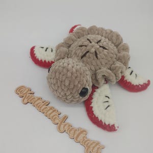 May include: A crocheted plush turtle toy with a brown body and red and white shell. The turtle is lying on its side with its head turned to the side. The toy is on a white background with a wooden sign that says "Knotalotta Acres Crafts".