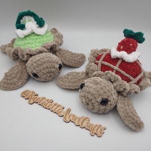 May include: Two crocheted turtle plushies with different colored shells. One turtle has a green shell with a white and green hat, and the other turtle has a red shell with a white and red hat. The turtles are both lying on a white surface.