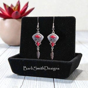 May include: A pair of silver-toned earrings with red gemstone accents and feather charms. The earrings have a geometric design with a central red stone and are displayed on a black velvet surface. The text "BarbSmithDesigns" is visible.