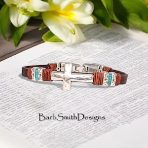May include: A brown leather bracelet with a silver cross pendant. The bracelet features turquoise accents and is secured with a silver clasp. The text "Barb Smith Designs" is visible.
