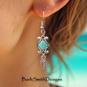 May include: A silver-toned earring with a turquoise square stone and a teardrop-shaped charm. The earring is suspended from a hook and features decorative scrollwork. The text "Barb Smith Designs" is visible at the bottom.