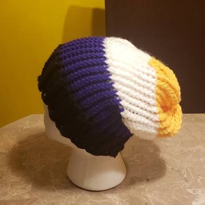 May include: A handmade beanie hat in a non-binary pride flag color scheme. The hat is crocheted with black, purple, white, and yellow yarn. The hat is displayed on a white mannequin head.