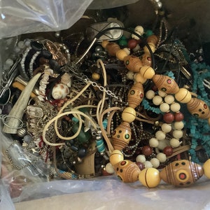 May include: A box overflowing with various jewelry pieces. The collection includes necklaces with wooden beads, turquoise beads, and shell accents. There are also bracelets, earrings, and other accessories in silver and gold tones. A variety of textures and styles are visible.
