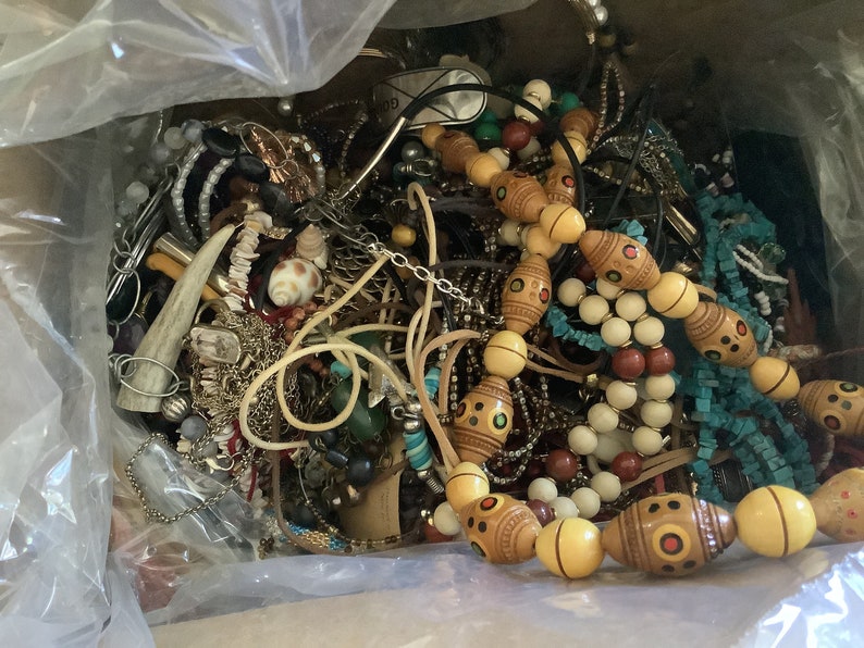 May include: A box overflowing with various jewelry pieces. The collection includes necklaces with wooden beads, turquoise beads, and shell accents. There are also bracelets, earrings, and other accessories in silver and gold tones. A variety of textures and styles are visible.
