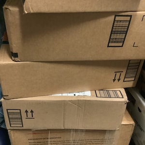 May include: A stack of brown cardboard boxes with black barcode labels and directional arrows. The boxes are of varying sizes and are stacked haphazardly. The boxes are in a storage area.