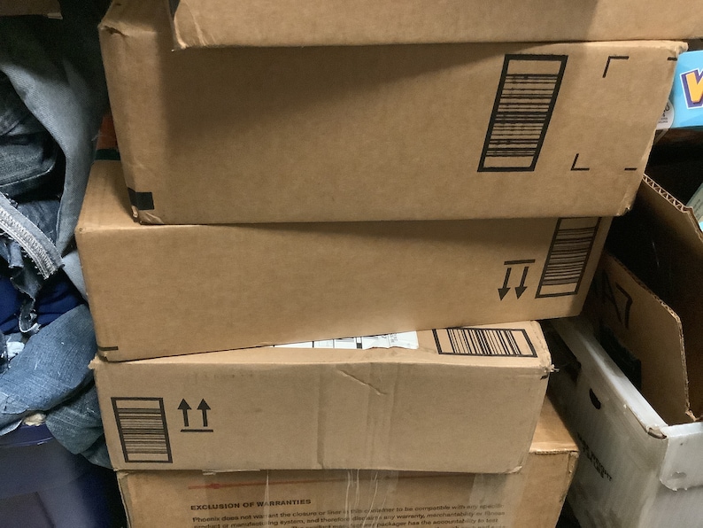May include: A stack of brown cardboard boxes with black barcode labels and directional arrows. The boxes are of varying sizes and are stacked haphazardly. The boxes are in a storage area.
