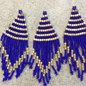May include: Three pairs of blue and gold beaded earrings. The earrings are long and dangle, with a triangular shape at the top and a fringe of beads at the bottom.