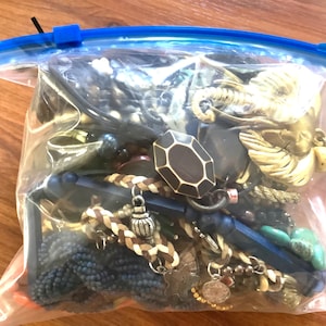 Peut inclure: Un sac en plastique transparent rempli de divers bijoux. Le sac contient des colliers, des bracelets et d'autres accessoires dans les tons de noir, bleu, marron et or. Certains articles ont des formes géométriques et des détails tressés.