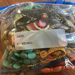 May include: A clear plastic bag filled with various jewelry pieces. The jewelry includes necklaces, bracelets, and earrings in a variety of colors, including turquoise, gold, red, and black. The bag has a blue zipper and a label that says "DATE" and "CONTENTS".