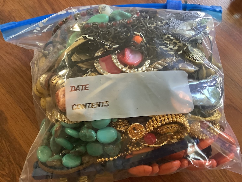 May include: A clear plastic bag filled with various jewelry pieces. The jewelry includes necklaces, bracelets, and earrings in a variety of colors, including turquoise, gold, red, and black. The bag has a blue zipper and a label that says "DATE" and "CONTENTS".