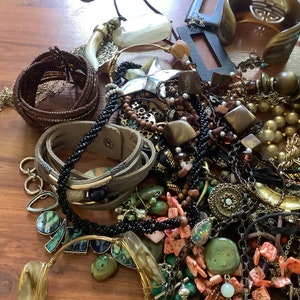 May include: A collection of various jewelry pieces, including necklaces, bracelets, and earrings. The assortment features a mix of materials, such as beads, leather, metal, and gemstones, in shades of brown, gold, green, and black. A star-shaped pendant is also visible.
