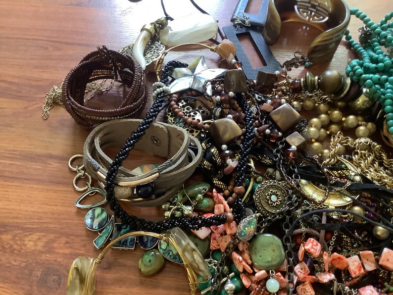 May include: A collection of various jewelry pieces, including necklaces, bracelets, and earrings. The assortment features a mix of materials, such as beads, leather, metal, and gemstones, in shades of brown, gold, green, and black. A star-shaped pendant is also visible.