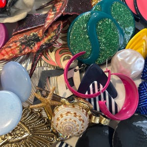 Könnte beinhalten: Eine Sammlung von bunten und einzigartigen Ohrringen, darunter ein Paar große grüne Glitzerohrringe, ein Paar pink-schwarz gestreifte Ohrringe und ein Paar schwarz-weiß gestreifte Ohrringe. Es gibt auch mehrere andere Ohrringe, darunter ein Paar blau-weiße Ohrringe, ein Paar goldene Ohrringe und ein Paar weiße Perlenohrringe.