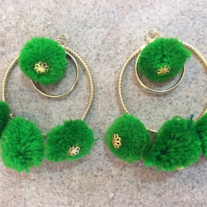 May include: A pair of green pom-pom earrings with gold hoops. Each earring has three green pom-poms and a gold circle with a small gold flower design.