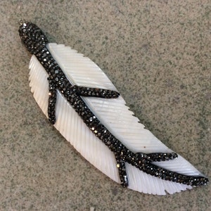 Peut inclure: Un pendentif en nacre blanche en forme de plume avec un motif de branche en strass noirs.