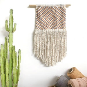 以下が含まれることがあります： ピンク、オレンジ、ブラウンの色合いの幾何学模様が織り込まれた壁掛け。フリンジの縁取りがあり、木製の棒に掛けられています。