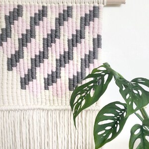 Peut inclure: Une tapisserie murale tiss&eacute;e rose, blanche et grise avec une bordure en franges. La tapisserie est faite de fil et pr&eacute;sente un motif g&eacute;om&eacute;trique.