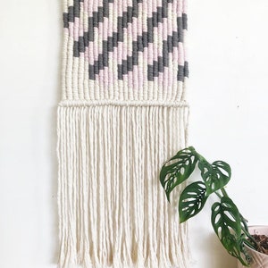Peut inclure: Une tapisserie murale en macram&eacute; de couleur rose, grise et blanche avec un motif g&eacute;om&eacute;trique et une longue frange. La tapisserie est mont&eacute;e sur une baguette en bois.