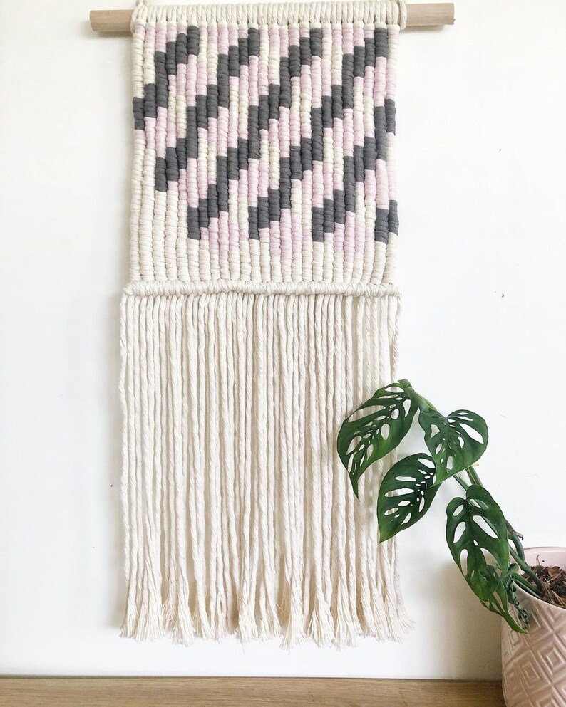 Peut inclure: Une tapisserie murale en macram&eacute; de couleur rose, grise et blanche avec un motif g&eacute;om&eacute;trique et une longue frange. La tapisserie est mont&eacute;e sur une baguette en bois.