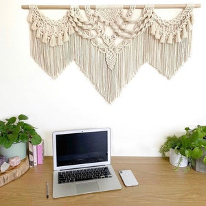 Op de afbeelding: Een witte macramé wandhanger met een geometrisch ontwerp hangt boven een houten bureau met een laptop, een telefoon en twee potplanten.