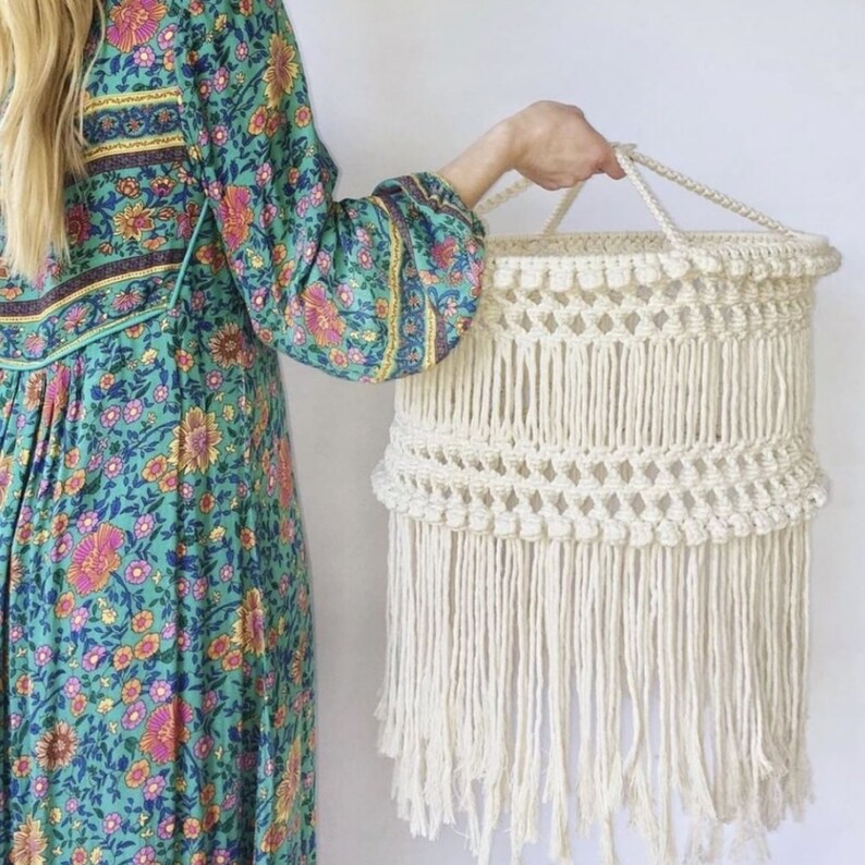 May include: A white macrame lampshade with a fringe bottom. The lampshade is hanging from a loop at the top.