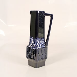 Puede incluir: Una jarra de cerámica azul oscuro con un diseño floral blanco y patrones geométricos. La jarra tiene una base cuadrada y un asa.