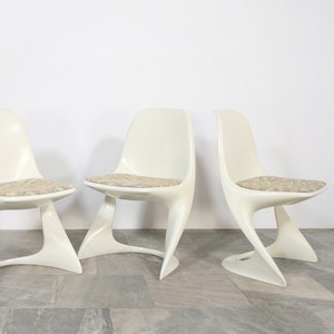 May include: Set of four white molded plastic chairs with a unique, curved design. Each chair has a beige patterned cushion.