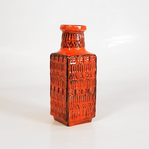 Puede incluir: Jarrón de cerámica naranja con un patrón geométrico. El jarrón tiene una base cuadrada y un cuello estrecho.