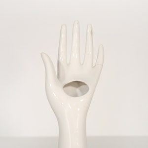 May include: White ceramic hand ring holder. The hand is outstretched with the fingers spread apart. There is a hole in the palm of the hand for holding a ring.