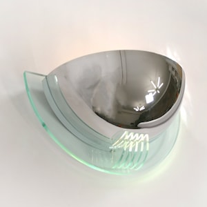 Könnte beinhalten: Eine Wandleuchte aus Chrom und Klarglas mit einem abgerundeten, reflektierenden Schirm. Die Leuchte hat ein modernes Design und ist ideal, um jedem Raum einen Hauch von Eleganz zu verleihen.