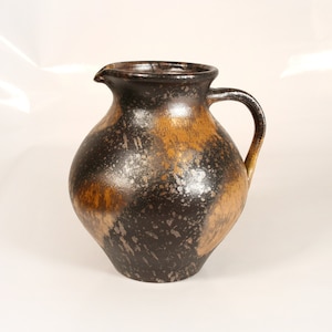 Puede incluir: Una jarra de cerámica marrón y negra con un esmalte moteado. La jarra tiene un cuerpo redondeado y una sola asa.