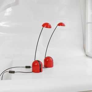 May include: Two red and black desk lamps with flexible arms and round shades. The lamps are on a white surface.